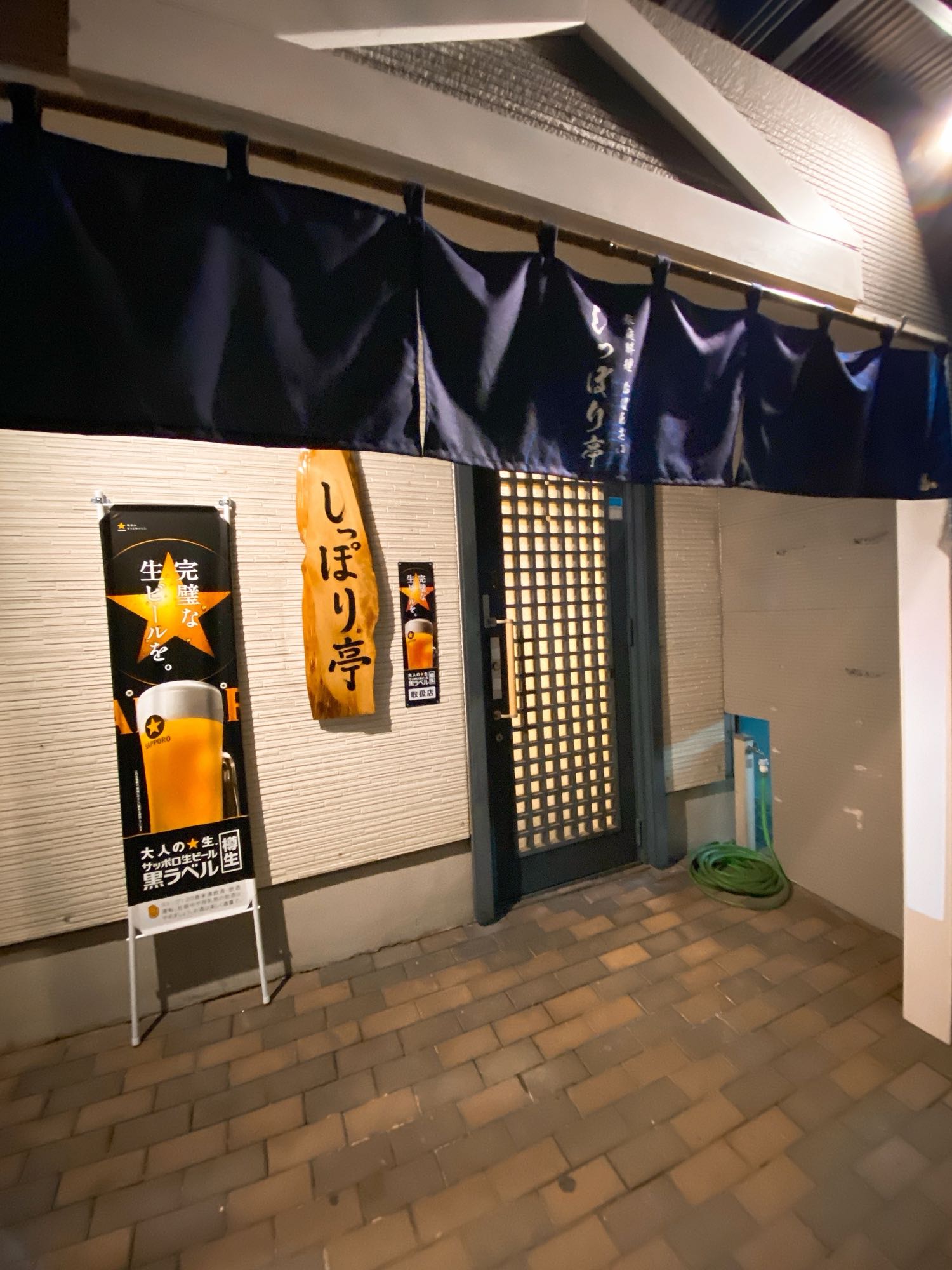 夜のしっぽり亭の入口。紺の暖簾とサッポロ黒ラベル(生)の看板
